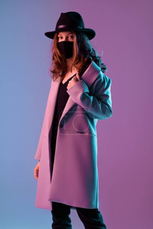 Téléchargez les photos : Portrait d'une jeune femme branchée en manteau, chapeau et masque de protection en néon violet. - en image libre de droit