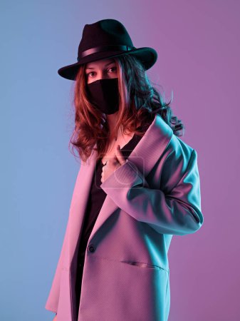 Photo for Portrait of a fashionable young woman in a coat, hat and protective mask in purple neon lighting. - Royalty Free Image