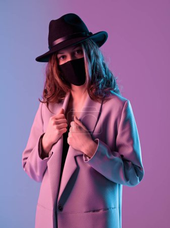 Téléchargez les photos : Surpris jeune femme à la mode dans un manteau, chapeau et masque de protection dans l'éclairage au néon violet. - en image libre de droit