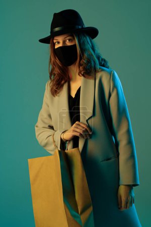 Téléchargez les photos : Portrait d'une jeune femme branchée en manteau, chapeau et masque de protection avec un sac à provisions en papier sur fond vert. - en image libre de droit