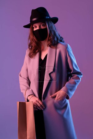Téléchargez les photos : Portrait d'une jeune femme branchée vêtue d'un manteau, d'un chapeau et d'un masque de protection avec un sac à provisions en papier en néon violet. - en image libre de droit