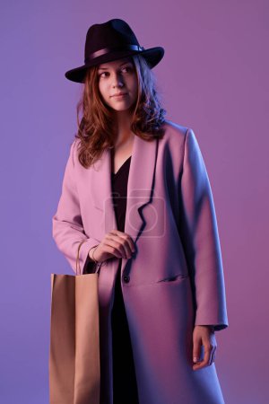 Foto de Retrato de una joven de moda en un abrigo y sombrero con una bolsa de compras de papel en iluminación de neón púrpura. - Imagen libre de derechos