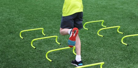 Photo for Young teenage boy jumping on one leg over six inch mini hurdles on a turf field during summer camp. - Royalty Free Image