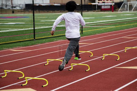 Photo for Rear view of a high school boy running the wicket drill on a red track over yellow mini hurdles during track practice. - Royalty Free Image
