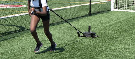 Foto de Una chica de secundaria está tirando de un trineo ponderado en un campo de césped durante la práctica de atletismo. - Imagen libre de derechos