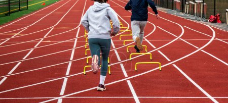 Photo for Rear view of two runners running over six inch yellow mini hurdles in lanes on a track at practice. - Royalty Free Image