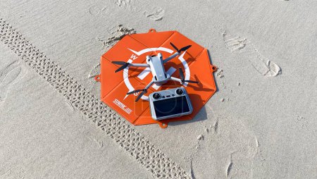 Foto de Fire island, Nueva York, EE.UU. - 20 de febrero de 2023: DJI mini pro 3 drone en una plataforma de despegue naranja con su controlador en la arena en la playa. - Imagen libre de derechos