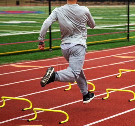 Photo for Rear view of a high school boy is running fast over six inch plastic yellow hurdles in lane on a track. - Royalty Free Image