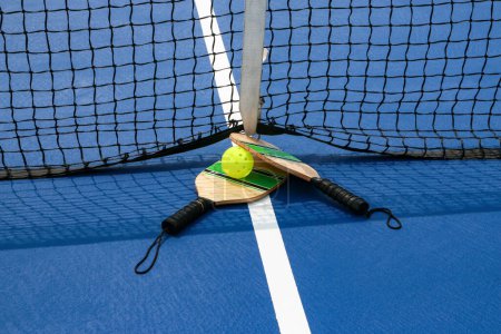 Foto de Primer plano de las paletas de pickleball y bola por la red en una cancha azul de cerca. - Imagen libre de derechos