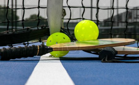 Foto de Close up of two pickleball rackets and two yellow whiffle balls next to the net on a tennis court. - Imagen libre de derechos