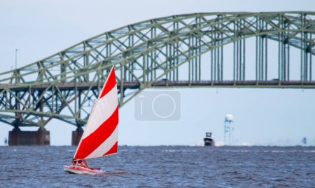 Front view of a close up of one red and white sunfish sailboat sailing on the Great South Bay with the bridge close in the background.