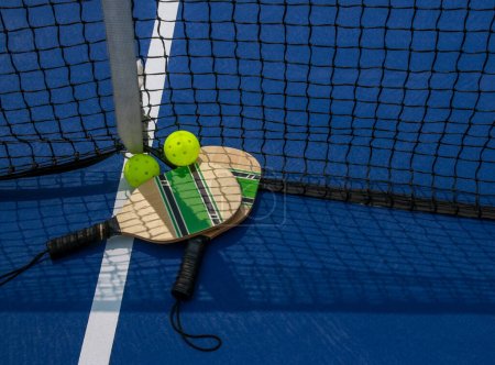 Two pickleball paddles and two yello whiffle balls on a court by the net close up
