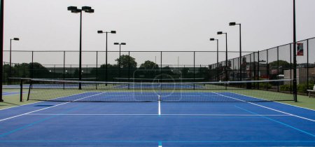 Photo for Front view of blue and green tennis courts with lights surrounded by tall black fencing. - Royalty Free Image