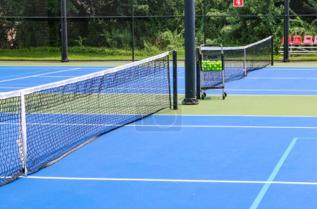 Foto de Vista lateral de una pista de tenis azul y verde con un carro lleno de pelotas de tenis encerradas con esgrima negra. - Imagen libre de derechos
