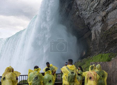 Niagara Falls, Ontario, Canada - 31 July 2023: Rear view of tourists taking pictures of the falls wearing yellow raingear on a viewing platform under the falls that are on the journey behind the falls tour.