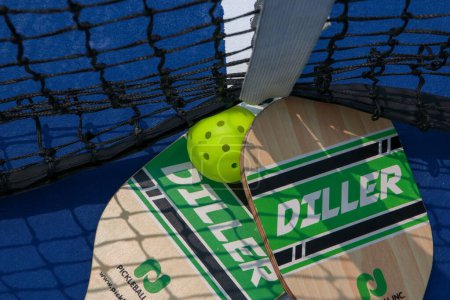 Foto de West Islip, Nueva York, EE.UU. - 17 de julio de 2023: Mirando hacia abajo a dos raquetas de pickleball en la red de tenis con un whiffle ball de cerca. - Imagen libre de derechos