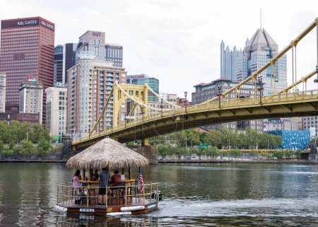 Foto de Pittsburgh, Pensilvania, EE.UU. - 6 de agosto de 2023: Un barco Tiki flotante bar con techo de paja en un río con un puente en el fondo en el río Allegany en Pittsburg. - Imagen libre de derechos