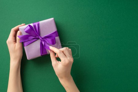 Photo for New Year concept. First person top view photo of young woman's hands unpacking lilac giftbox with violet ribbon bow on isolated green background with empty space - Royalty Free Image
