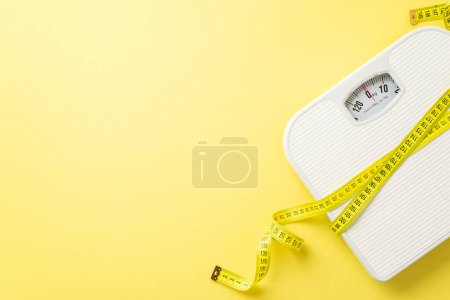 Slimming concept. Top view photo of measuring tape wrapped around scales on isolated pastel yellow background with copyspace
