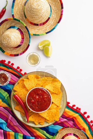Photo for Spice things up with Cinco de Mayo-inspired vertical flat lay! Mexican hat, poncho maracas, tequila shots, lime, chili peppers, nacho chips salsa arranged on white backdrop space for your message - Royalty Free Image