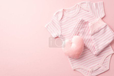 Babykonzept. Foto von oben von Säuglingskleidung rosa Anzughose und flauschiges herzförmiges Spielzeug auf isoliertem pastellrosa Hintergrund mit leerem Raum