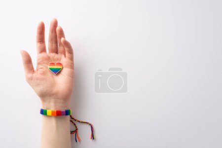 A first person top view photo of a female hand in a rainbow-colored bracelet holding a heart-shaped pin badge on a white background with space for text or advertising, celebrating LGBTQ History Month