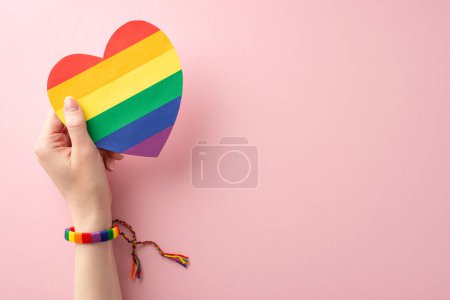Téléchargez les photos : Thème du Mois de l'histoire LGBT : photo à la première personne de la main de la jeune femme en bracelet symbolique tenant une carte coeur arc-en-ciel sur fond rose pastel avec espace pour le texte ou la publicité - en image libre de droit