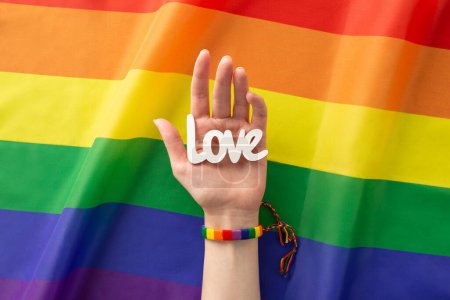 Téléchargez les photos : Une vue de haut en bas à la première personne de la main d'une femme arborant un bracelet arc-en-ciel et tenant le mot "amour" sur sa paume sur un fond ondulé de drapeau LGBTQ offre beaucoup d'espace pour la publicité - en image libre de droit