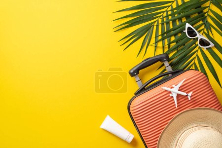 Tropical journey concept. Above view photo of orange suitcase with airplane model and straw hat on it surrounded by palm leaves and sunglasses on isolated bright yellow background with copy-space