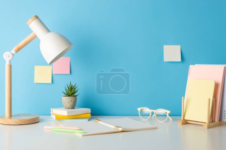 Photo for Academic essentials, such as books holder, copybook, glasses, and lamp neatly arranged on desk, captured in side view photo against blue wall with sticky notes. Perfect for text or promotional content - Royalty Free Image
