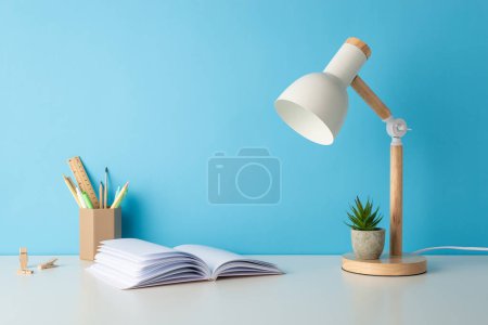 Photo for Side view photo of desk equipped with school supplies: pens holder, pencils, ruler, pins, open notebook, lamp, and small flowerpot. Blue wall background, excellent for text or advert usage - Royalty Free Image