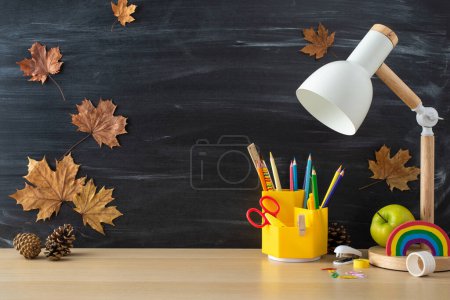 Photo for Elevate your study environment with this student's workplace. Side view photo highlighting lamp, stationery on a wooden desk on blackboard isolated background and copy-space for text or advertising - Royalty Free Image