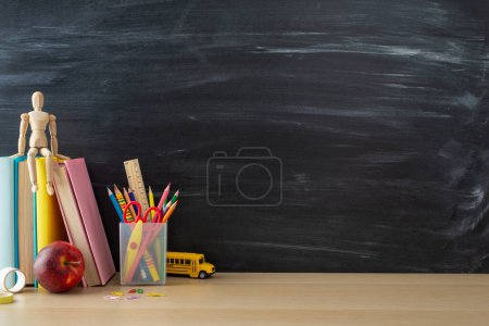 Photo for School supplies layout. Side view photo of desk setup with pencils organizer, ruler, books, red apple, mannequin body, and more on chalkboard background. Great for educational content or advertising - Royalty Free Image
