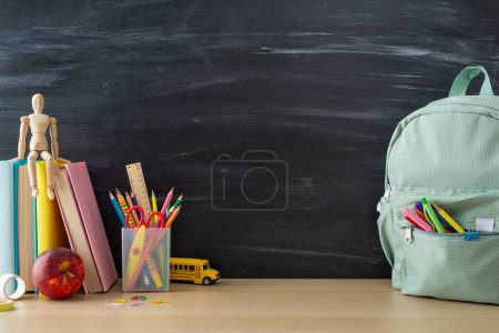 Photo for Unlock your creativity - side view photo of desk decorated with pencils holder, books, clips, apple. Backpack, mannequin body, and toy school bus enhance atmosphere. Chalkboard backdrop await ideas - Royalty Free Image