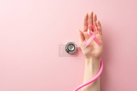 Photo for Honor Breast Cancer Awareness Month with this top view image of female hand holding pink ribbon and stethoscope on pastel pink isolated background with copyspace available for text or ads - Royalty Free Image