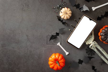 Make your Halloween greetings stand out: first person top view of skeleton hand, holding smartphone, pumpkins, eerie spiders, spiderweb, bats, bones on dark grunge backdrop, space for text or promo