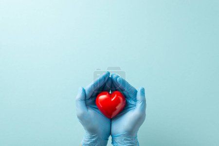 Photo for Heart disease defense strategy with this captivating birds-eye top scene displaying gloved hands cradling a heart on pale blue background with copy-space available for text or promotional use - Royalty Free Image
