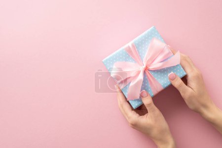 Téléchargez les photos : Vue de haut en bas de la première personne des mains féminines, caressant une boîte-cadeau bleu pastel à pois et un délicat ruban rose noeud, sur un fond rose pastel, laissant place à votre message - en image libre de droit
