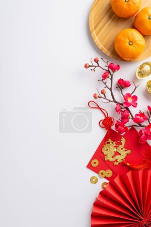 Explore this Chinese New Year scene: top-view vertical shot of fan, ancient coins, lucky sycee, red Hong Bao, dragon charm hanging, tangerine-filled plate, sakura, white backdrop with space for text