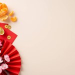 Ignite the New Year festivities with this charming composition. Top-down view of red fan, feng shui adornments, lucky coins, red envelopes, peeled tangerines on neutral background with space for text