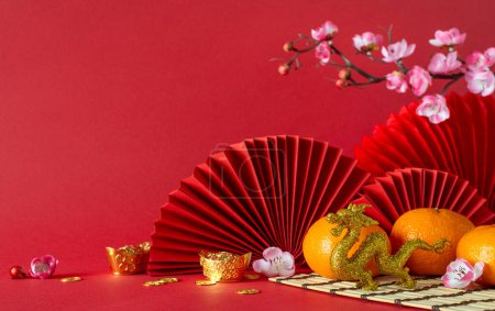 Photo for Traditional New Year tableau: side view feng shui elements, gold coins, dragon ornament, tangerines, sakura flowers, and fans arranged on red surface, providing a perfect canvas for text or promotions - Royalty Free Image