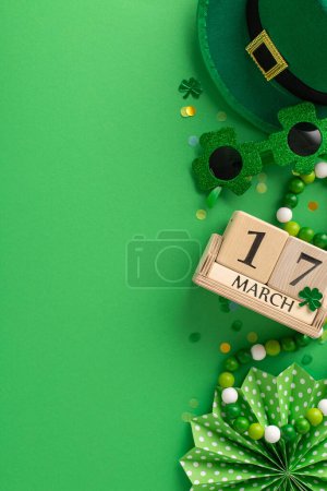 Photo for March 17th celebration display: vertical top view wooden calendar with the date, leprechaun's hat, party glasses, fan, confetti, and beads arranged on a green background for text placement - Royalty Free Image