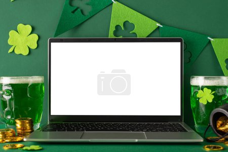 Photo for St. Patrick's Day theme: side view photo of laptop open for virtual celebrations, frothy beer mugs, scattered gold coins, pot, and trefoil flag streamers on a vibrant green surface with space for text - Royalty Free Image
