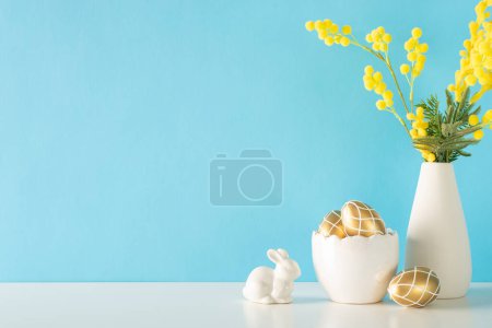Téléchargez les photos : Scène de cuisine de vacances, présentant une vue latérale d'un comptoir avec un bol en porcelaine d'œufs peints en or, une statuette de lapin et un pichet de tulipes sur un fond de mur bleu clair pour le texte - en image libre de droit