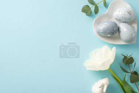 Spring celebration display: Top view dove grey eggs in an heart shaped platter, bunny ornament, white tulip, and green eucalyptus on a sky-blue backdrop, space for text