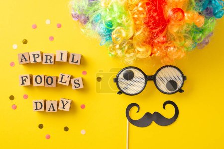 Schnappschuss von Holzbuchstaben mit der Aufschrift "April Fool 's Day", Konfetti mit Party-Accessoires wie Clownsperücke, Brille und Schnurrbart, die ein humorvolles Gesicht auf einer leuchtend gelben Oberfläche erzeugen