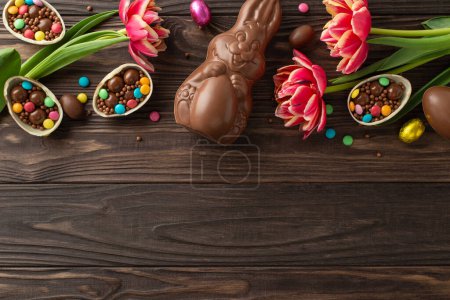 Delightful Easter treat scene. Overhead shot of broken chocolate shells, brimming with vibrant sweets, a chocolate rabbit, and bright tulips on a wooden backdrop, leaving space for text or ads