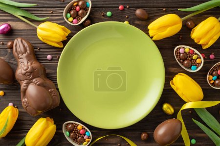 Easter celebration sweets scene: from top view, see an empty green plate, chocolate eggs spilling candies, a chocolate rabbit, and tulips on a wooden table, with area for text or advertisement