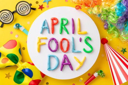 Foto von oben zeigt "April Fool 's Day" in Tonbuchstaben, arrangiert in rundem Rahmen, Comicbrille, Narrenkappe, bunte Krawatte, Gebläse und mehr, alles arrangiert auf leuchtend gelbem Hintergrund