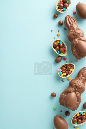 Adorable Easter ensemble idea. Vertical top view of cracked chocolate eggs, filled with colorful candies, a chocolate bunnies, sprinkles on a pastel blue background, placeholder for text or advert